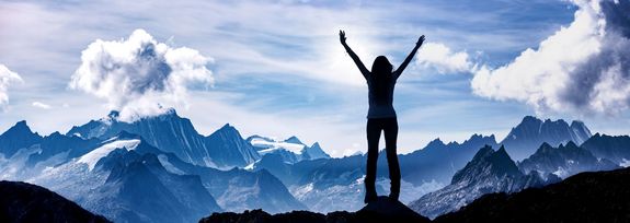 Die Silhouette einer Frau, die auf einem Berg steht, beide Arme in die Höhe streckt und in die Ferne auf einen strahlend blauen Himmel und weitere Bergkämme blickt